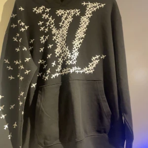 Svart hoodie från Louis Vuitton med flygplansmönster - Säljer en svart hoodie från Louis Vuitton med stort LV-tryck och flygplansmönster över bröstet och ärmen. Tröjan har huva och stor magficka framtill. Perfekt för dig som vill sticka ut med en exklusiv streetwear-look. Storlek XL men passar L