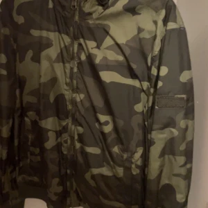 Woolrich jacka vändbar - Säljer en snygg vindjacka med camouflage-mönster och huva. Jackan har dragkedja framtill och är perfekt för dig som gillar streetwear-stil. Passar bra till vår och höst.