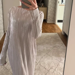 Silverfärgad långklänning med glittereffekt från soft rebels - Säljer en lång, glittrig silverklänning från soft rebels med lång ärm och rund halsringning. Klänningen har en rak passform och skimrande material som ger en festlig look. Perfekt för speciella tillfällen!