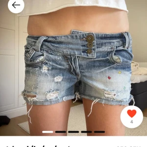SÖKER!!! - Säljer ett par blå jeansshorts med låg midja och flera slitningar samt fransar nertill. Shortsen har tre dekorativa knappar framtill och små färgglada broderade detaljer. Perfekta för sommaren!
