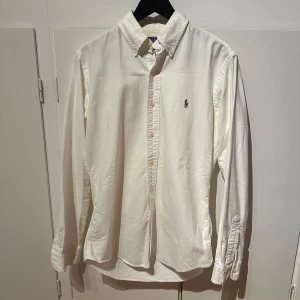 Vit skjorta från Polo Ralph Lauren - Klassisk vit skjorta från Polo Ralph Lauren med broderad logga på bröstet. Skjortan har lång ärm, knappar framtill och är i bomull. Perfekt för en clean och stilren look.
