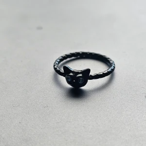 Svart ring med kattmotiv - Cool svart ring med ett kattmotiv framtill och blå detaljer som ögon. Ringen har en tvinnad design på bandet och är perfekt för dig som gillar unika accessoarer. Rostfritt stål, 16.9 mm