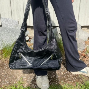 Svart handväska med dragkedjor - Säljer en svart handväska med två snedställda dragkedjor framtill. Väskan har en cool, lite glansig yta och bred axelrem. Perfekt för dig som vill ha en stilren men edgy accessoar till din outfit💘