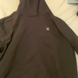 Svart hoodie från Calvin Klein Jeans - Svart hoodie från Calvin Klein Jeans med liten CK-logga på bröstet. Klassisk modell med huva och långa ärmar, perfekt för en avslappnad stil.