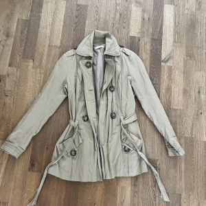 Beige trenchcoat med knytskärp - Säljer en beige trenchcoat med knytskärp i midjan och dekorativa knappar fram och bak. Jackan har klassisk krage, långa ärmar och fickor med lock. Perfekt för övergångssäsonger.