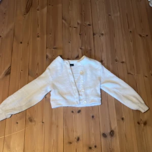 Vit fluffig kofta från Gina Tricot - Supermjuk och fluffig vit kofta från Gina Tricot. Modellen är croppad med långa ärmar och knäpps med vita knappar framtill. Perfekt för lager-på-lager och ger en mysig känsla till din outfit.