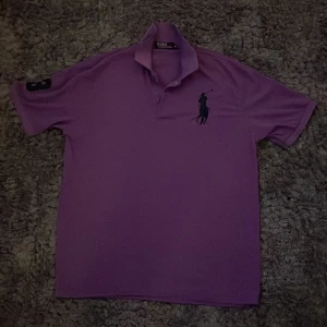 Lila pikétröja från Polo Ralph Lauren - Säljer en lila pikétröja från Polo Ralph Lauren med klassisk krage och knäppning framtill. Stor logga broderad på bröstet och siffra på ärmen. Perfekt för en stilren och sportig look.