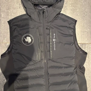 Svart dunväst från Sail Racing - Säljer en svart dunväst från Sail Racing med huva och dragkedja framtill. Västen har en bröstficka med dragkedja och logotyp på vänster sida. Använd två gånger och sitter normalt i storleken. 