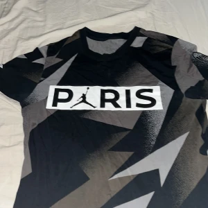 PSG x Jordan Paris Saint-Germain fotbollströja - Snygg svart och grå PSG x Jordan fotbollströja med grafiskt mönster och PARIS-tryck på bröstet. Har klubbmärke på ärmen och texten 'ICI C'EST PARIS'. Perfekt för dig som gillar streetwear och fotboll. Helt äkta man se från jorden logon.