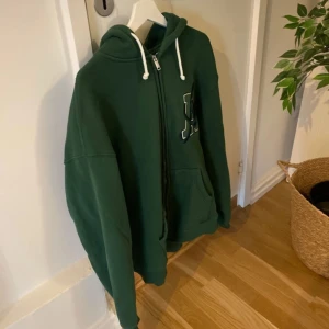 Grön hoodie med dragkedja från Axel Arigato - Mörkgrön hoodie från Axel Arigato med dragkedja framtill, vit snörning i huvan och stor ficka på magen. Stor bokstavsdetalj på bröstet. Perfekt för en avslappnad stil.