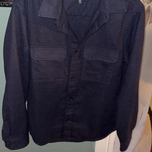 Mörkblå overshirt från H&M - Säljer en mörkblå overshirt från H&M med klassisk krage och två bröstfickor. Skjortan har långa ärmar och knäppning framtill. Perfekt att bära som lager över en t-shirt eller tröja.