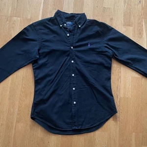 Svart skjorta från Polo Ralph Lauren - Klassisk skjorta i svart från Polo Ralph Lauren, med lila logga broderad på bröstet. Slim fit-modell i mjuk bomull med button-down-krage och vita knappar. Passar både till vardags och när du vill klä upp dig lite extra.