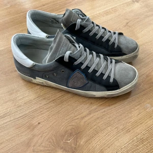 Philippe Model sneakers i grå mocka och skinn - Snygga Philippe Model sneakers i grå mocka och svart skinn med vita detaljer och vit sula. Skorna har snörning och en ikonisk sköld på sidan. Perfekta för en stilren och avslappnad look.