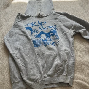 Grå hoodie med blått Mount Rap More-tryck - Säljer en grå hoodie med blått tryck på bröstet där det står 'Mount Rap More' och har motiv av fyra personer. Tröjan har huva och magficka. Defekt syns på bild 3 snöre går att sätta in men inget som finns i nu läget