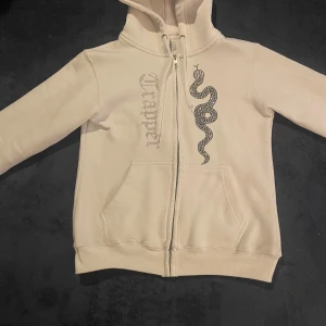 Vit hoodie från trapper - Säljer en vit trappee hoodie med dragkedja och huva. På framsidan finns en stor orm i svarta strasstenar och texten 'trapper' i glittriga stenar. Jackan har två fickor framtill och ribbade muddar vid ärmslut och nederkant.
