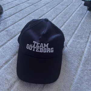Svart keps Team Göteborg - Svart keps med vit broderad text 'TEAM GÖTEBORG' framtill. Justerbar rem baktill för perfekt passform. Tillverkad i polyester och har klassisk böjd skärm. Perfekt för dig som vill visa Göteborg-stolthet.