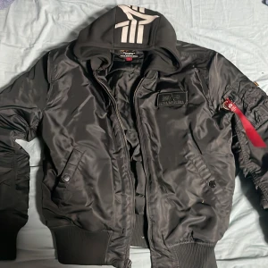 Svart bomberjacka från Alpha Industries - Säljer en svart bomberjacka från Alpha Industries i storlek L. Jackan har dragkedja, ribbade muddar och tydliga loggor både fram och bak. Klassisk modell med fickor och ikonisk röd tag på ärmen. Säljer för 1500kr nypris 2500kr