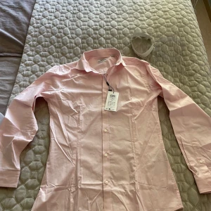 Ljusrosa skjorta från Jack & Jones - Säljer en stilren ljusrosa skjorta från Jack & Jones. Skjortan har klassisk krage, långa ärmar och knäppning framtill. Perfekt för dig som vill ha en fräsch och enkel look. Märkt med 'Premium' på etiketten.