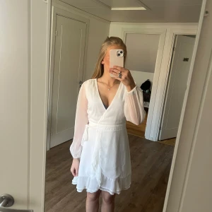 Student klänning - Säljer en vit omlottklänning från Bubbleroom med volangdetaljer nertill, använd 1 gång men ser ut som ny. Klänningen har v-ringning och knytband i midjan för en smickrande passform. Perfekt för sommarens tillfällen och framförallt till skolavslutning eller student. Midjeviddenär ungefär 65-70cm. Bystvidden är 82cm!