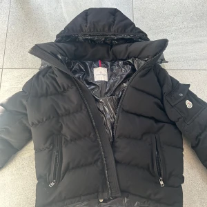 Svart pufferjacka från Moncler - Säljer en svart pufferjacka från Moncler med huva och dragkedja. Jackan har två fickor framtill med dragkedja och en ficka med knapp på ärmen där Moncler-loggan sitter. Insidan har ett serietidningsmönster.