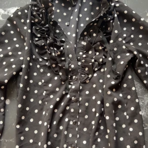 Svart prickig blus med volang - Säljer en svart blus/body med vita prickar och volangdetaljer längs knäppningen. Blusen har långa ärmar och knappar framtill. Perfekt för dig som gillar en lekfull och trendig stil. Är i strl XL men passar även i S och uppåt beroende på hur man vill att den ska sitta