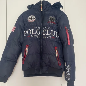 Blå dunjacka från Santoz Polo Club - Mörkblå dunjacka med huva från Santoz Polo Club. Jackan har broderade och tryckta loggor fram, bak och på ärmen samt dragkedjor med röda detaljer. Fodrad och med ribbade muddar vid ärmslut och nederkant. Finns märke som inte går bort 💕 storlek S