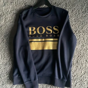 Mörkblå sweatshirt från Hugo Boss - Snygg mörkblå sweatshirt st M passar som S från Hugo Boss med stort guldigt BOSS-tryck på bröstet. Tröjan har rund halsringning, långa ärmar och ribbade muddar vid ärmslut och nederkant. Perfekt för en stilren och avslappnad look.
