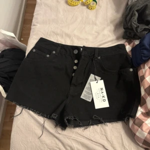 Svarta jeansshorts från NA-KD - Svarta jeansshorts från NA-KD med råa, fransiga kanter och klassisk femficksdesign. Knappar framtill och hög midja. Perfekta för sommaren och enkla att matcha med olika toppar.