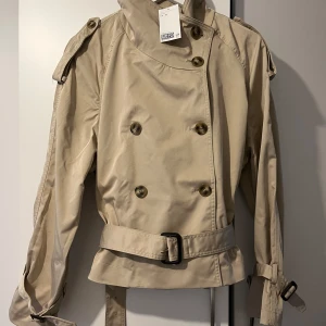 Beige trenchcoat med bälte - Kort beige trenchcoat med dubbelknäppning, axelklaffar och bälte i midjan. Jackan har långa ärmar med spännen vid ärmsluten och klassisk krage. Perfekt för övergångssäsonger och ger en stilren look.