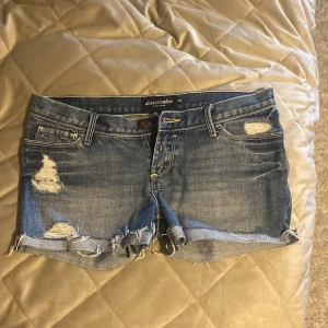 Blå jeansshorts från Abercrombie - Säljer ett par blå jeansshorts från Abercrombie med slitna detaljer och fransig nederkant. Klassisk femficksmodell med låg midja och råa kanter. Perfekta för en avslappnad stil. Ungefär 40 cm rakt över