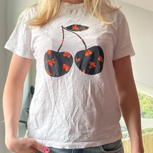 Vit t-shirt med körsbärstryck från Ganni - Vit t-shirt från Ganni med ett stort svart körsbärstryck framtill, dekorerat med små röda blommor. Klassisk passform och rund halsringning. Perfekt för en avslappnad och trendig look.