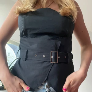 Svart axelbandslös topp med bälte - Snygg svart axelbandslös topp med markerad midja och tillhörande bälte. Toppen har dragkedja bak och en asymmetrisk nederkant framtill. Perfekt att styla med jeans eller kjol. från zara