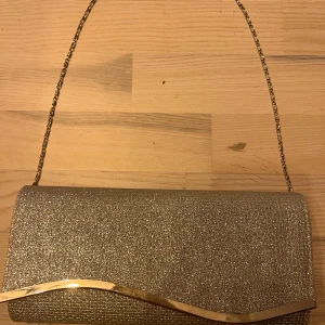 Glittrig kuvertväska med gulddetalj - Säljer en glittrig kuvertväska i silver med kedjerem och snygg vågig gulddetalj framtill. Perfekt för att lyfta din outfit med lite extra bling. Magnetlås och ett litet innerfack för småsaker. Använd en gång. 