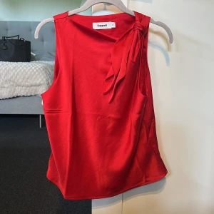 Röd ärmlös topp med rosett från Kappahl - Säljer en snygg röd ärmlös topp från Kappahl i glansigt material. Toppen har en dekorativ rosett vid axeln och en enkel, elegant design. Perfekt att styla med jeans eller kjol för en trendig look.
