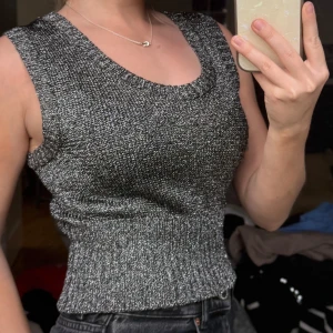 Glittrig stickad ärmlös topp - Säljer en glittrig stickad topp i svart och silver med breda axelband och rund halsringning. Toppen är tight och har en ribbad midja som ger en snygg passform. Perfekt för att lyfta vilken outfit som helst!