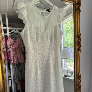 Vit spetsklänning med prickig mesh - Säljer en söt vit kortklänning med spets och prickig mesh upptill. Klänningen har ärmlös design, rund halsringning och en liten öppning i ryggen. Perfekt för sommarens tillfällen eller studentfirande. Klänningen är använd ett fåtal gånger! 