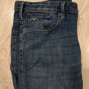 Jeans från Emporio Armani - Säljer ett par mörkblå jeans från Emporio Armani med klassisk femficksdesign. Byxorna har raka ben, normal passform och detaljer som metallogo på fickan och läderpatch bak i midjan. 