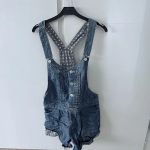 Jeansshorts hängselshorts med stjärnmönstrat foder - Säljer ett par blå jeansshorts med hängslen och knappar framtill. Shortsen har flera fickor och ett grått innerfoder med vita stjärnor. Perfekta för en avslappnad stil under varmare dagar.