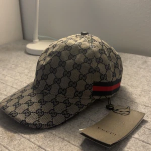 Beige gucci keps - Snygg keps fràn Gucci med klassiskt mönster i beige och brunt. Kepsen har en röd och grön randdetalj pà sidan. Perfekt för att ge din outfit en lyxig touch. Storlek "M”