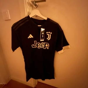 Svart Juventus fotbollströja från Adidas - Snygg svart Juventus fotbollströja från Adidas med vita detaljer och klubbens emblem på bröstet. Tröjan har korta ärmar och den ikoniska Jeep-loggan på framsidan. Perfekt för alla Juve-fans! ⚽️