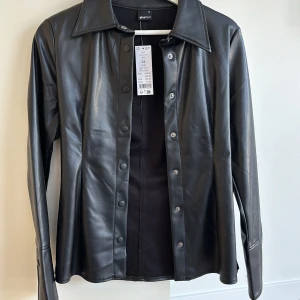 Svart PU-skjorta från Gina Tricot - Snygg svart skjorta i PU-material från Gina Tricot. Skjortan har långa ärmar och knappar framtill. Perfekt för en stilren look.