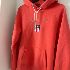 Hoodie från GANT - Säljer en snygg röd hoodie från GANT med broderad logga och emblem på bröstet. Tröjan har en klassisk känguruficka och justerbar huva med vita snören. Perfekt för en avslappnad stil. Nypris: 1600kr