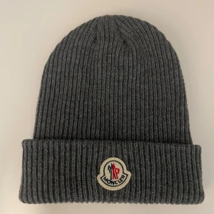 Grå ribbad mössa från Moncler - Snygg grå ribbad mössa från Moncler med broderad logotyp framtill. Perfekt för kyliga dagar och ger en stilren look. Passar både till vardags och mer uppklädda tillfällen.       Priset kan alltid diskuteras vid en snabb och smidig affär