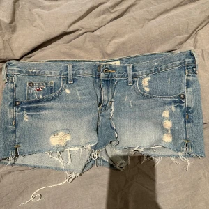 Blå jeansshorts från Hollister - Säljer ett par coola blå jeansshorts från Hollister med slitna detaljer och fransar. Shortsen har en klassisk femficksdesign och är perfekta för en avslappnad stil. De är tillverkade i 100% bomull och har en bekväm passform.