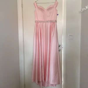 Ljusrosa långklänning med pärldetaljer - Elegant ljusrosa långklänning i satin med vacker pärldetalj i midjan. Klänningen har axelbandslös design och en flytande kjol som ger en romantisk känsla. Perfekt för speciella tillfällen. Är i storlek L men skulle kunna passa någon i storlek M med större byst. Ny pris 3500kr