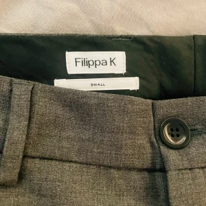 Filippa K Grå, lågmidjade kostymbyxor  - Snygga grå, lågmidjade kostymbyxor från Filippa K i en klassisk design. Byxorna har en rak passform med knapp och dragkedja framtill. Perfekta för en stilren look.