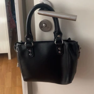 Svart handväska från NLY Accessories - Snygg svart handväska från NLY Accessories i syntetmaterial. Väskan har en praktisk dragkedja på framsidan och två handtag för enkel bärning. Perfekt för att hålla dina saker organiserade och lättillgängliga.