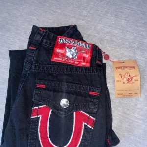 Svarta jeans från True Religion - Snygga svarta jeans från True Religion med ikonisk broderad logga i rött och vitt på bakfickorna. Jeansen har en klassisk femficksdesign och knappar i silver. Perfekta för en stilren look. helt nya har aldrig använt 