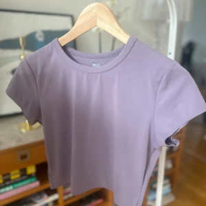 Lila t-shirt från Uniqlo - Säljer en stilren lila t-shirt från Uniqlo i storlek S. T-shirten är kortärmad och tillverkad av en mjuk polyesterblandning med elastan för extra stretch. Perfekt för en avslappnad look.