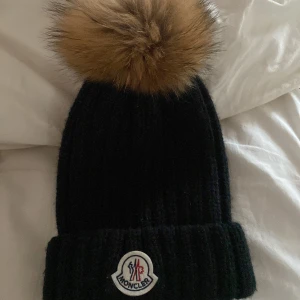 Svart stickad mössa från Moncler - Snygg svart stickad mössa från Moncler med en stor pälsboll på toppen. Mössan har en ribbad design och Moncler-loggan framtill. Perfekt för att hålla sig varm och stilren under kalla dagar.